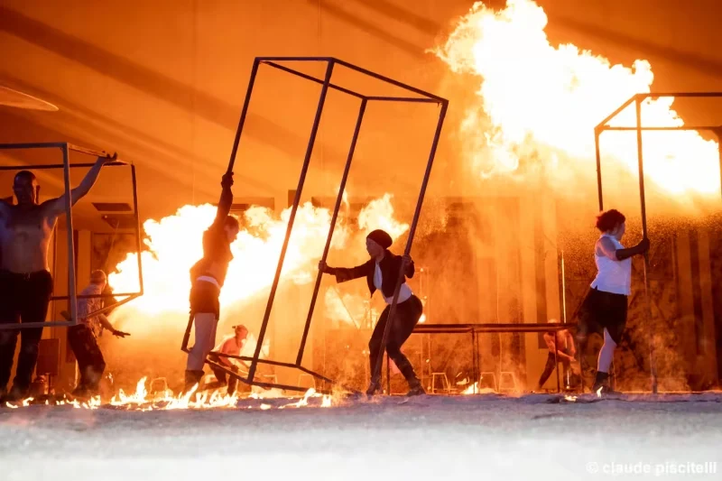 Scène du spectacle Merveilles avec plusieurs artistes manipulant des cadres métalliques devant un grand mur de flammes.