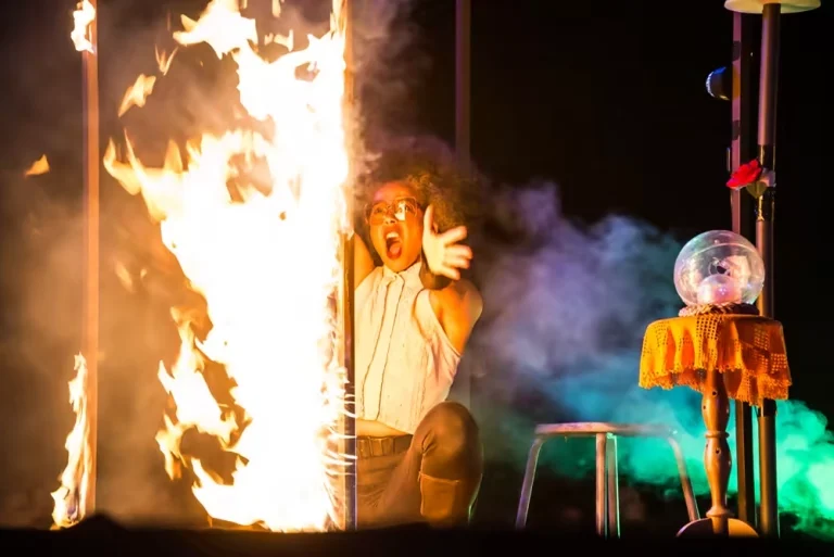 Artiste en costume clair réagissant devant une colonne de feu, avec décor théâtral du spectacle Merveilles.