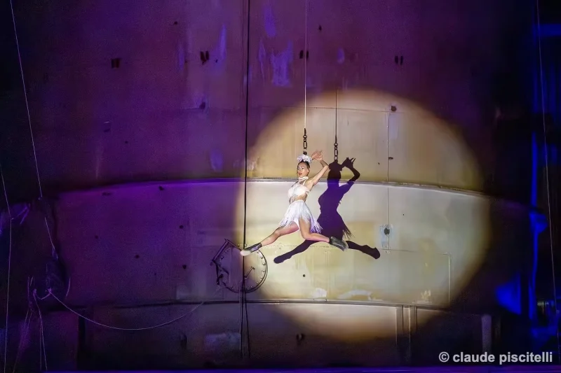 Acrobate suspendu devant une façade éclairée par un cercle de lumière pendant le spectacle Y’a d’la joie.