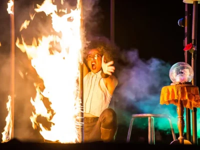 Artiste en costume clair réagissant devant une colonne de feu, avec décor théâtral du spectacle Merveilles.